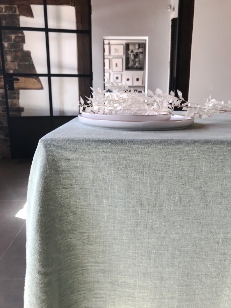 Puede incluir: Un mantel de lino verde claro con un plato blanco y ramas blancas secas en la parte superior.