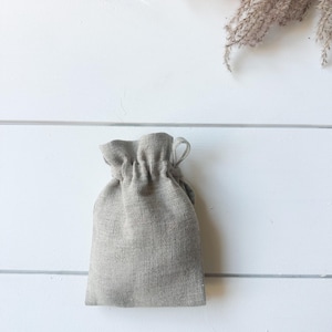 Puede incluir: Una peque&ntilde;a bolsa con cord&oacute;n de color gris claro, hecha de tela texturizada. La bolsa se cierra en la parte superior con un dise&ntilde;o fruncido. El fondo es una superficie de madera blanca con una ramita de hierba ornamental seca.