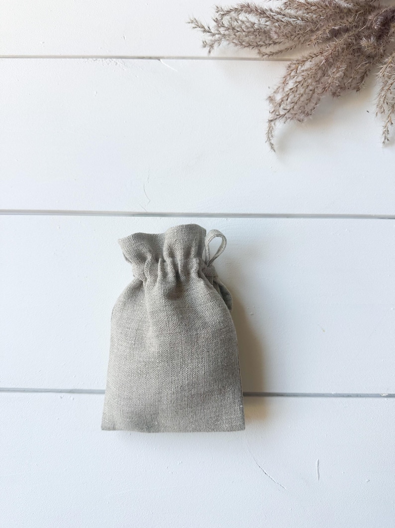 Puede incluir: Una peque&ntilde;a bolsa con cord&oacute;n de color gris claro, hecha de tela texturizada. La bolsa se cierra en la parte superior con un dise&ntilde;o fruncido. El fondo es una superficie de madera blanca con una ramita de hierba ornamental seca.