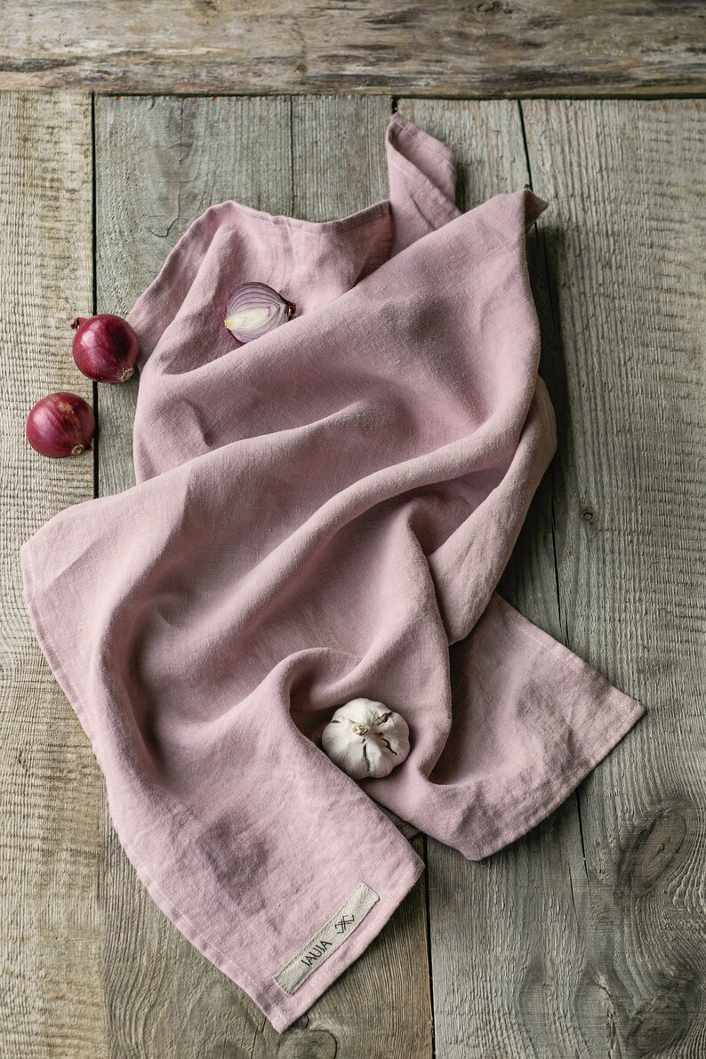 Op de afbeelding: Een roze linnen keukendoek met een wit label met de tekst "JAUJA" op een houten oppervlak. Er liggen drie rode uien en een teentje knoflook op de doek.