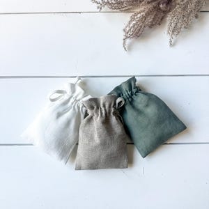 Puede incluir: Tres peque&ntilde;as bolsas con cord&oacute;n en blanco, gris y verde est&aacute;n dispuestas sobre una superficie de madera blanca. Las bolsas est&aacute;n hechas de una tela texturizada. Hierba ornamental seca en el fondo.