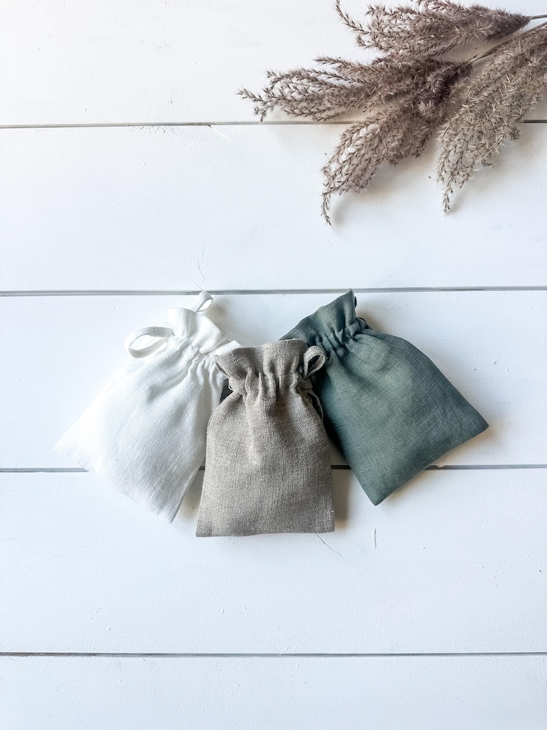 Puede incluir: Tres peque&ntilde;as bolsas con cord&oacute;n en blanco, gris y verde est&aacute;n dispuestas sobre una superficie de madera blanca. Las bolsas est&aacute;n hechas de una tela texturizada. Hierba ornamental seca en el fondo.