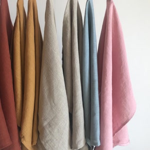 May include: Seven linen tea towels hanging on a white rod. The towels are in various colors, including rust, mustard yellow, natural linen, light blue, and pink.
