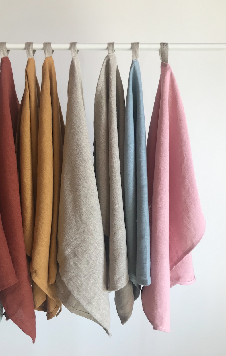 May include: Seven linen tea towels hanging on a white rod. The towels are in various colors, including rust, mustard yellow, natural linen, light blue, and pink.