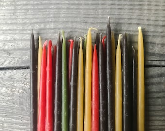 May include: A collection of thin, taper candles in various colors, including red, black, green, and yellow. The candles are arranged in a row on a wooden surface.