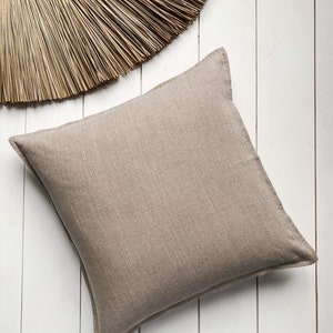 Pode incluir: Uma almofada de linho de cor castanho claro, de forma quadrada, com uma textura subtil. A almofada está colocada sobre uma superfície de madeira branca.