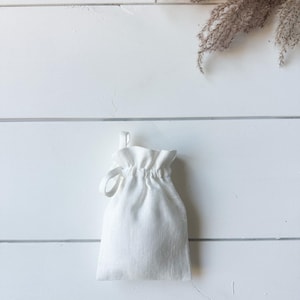Puede incluir: Una peque&ntilde;a bolsa blanca con cord&oacute;n hecha de tela texturizada. La bolsa est&aacute; fruncida en la parte superior con un cierre de cord&oacute;n en bucle. El fondo es una superficie de madera blanca con una ramita de plantas secas.