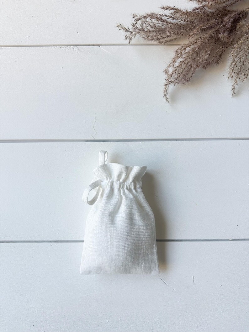 Puede incluir: Una peque&ntilde;a bolsa blanca con cord&oacute;n hecha de tela texturizada. La bolsa est&aacute; fruncida en la parte superior con un cierre de cord&oacute;n en bucle. El fondo es una superficie de madera blanca con una ramita de plantas secas.