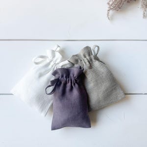 Puede incluir: Cuatro peque&ntilde;as bolsas con cord&oacute;n en blanco, gris y morado est&aacute;n dispuestas sobre una superficie de madera blanca. Las bolsas est&aacute;n hechas de lino o un tejido similar. Las bolsas se recogen en la parte superior con un cierre de cord&oacute;n.