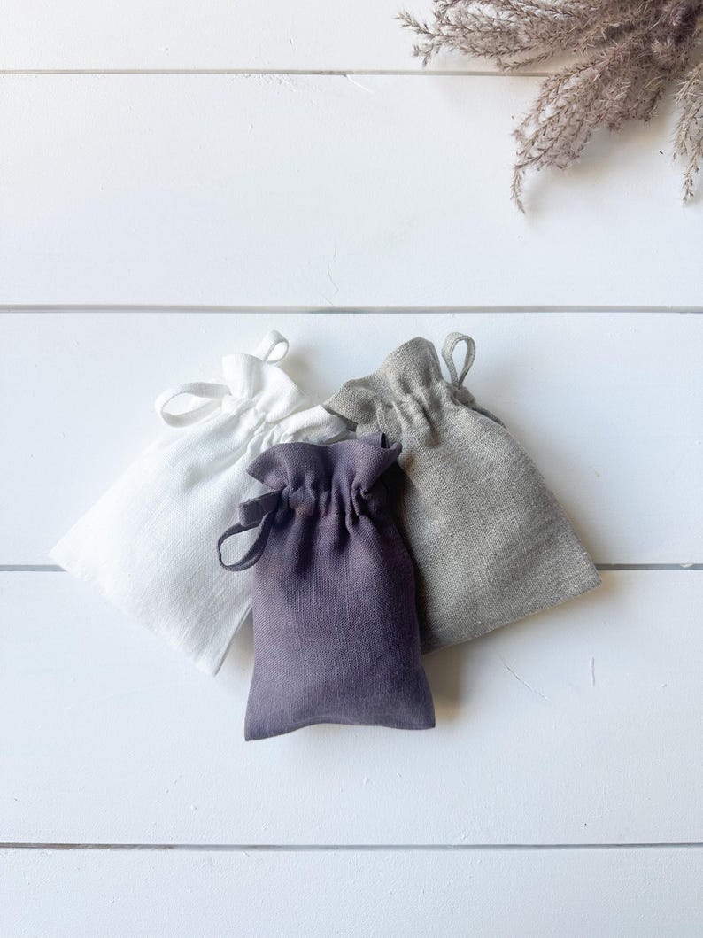 Puede incluir: Cuatro peque&ntilde;as bolsas con cord&oacute;n en blanco, gris y morado est&aacute;n dispuestas sobre una superficie de madera blanca. Las bolsas est&aacute;n hechas de lino o un tejido similar. Las bolsas se recogen en la parte superior con un cierre de cord&oacute;n.