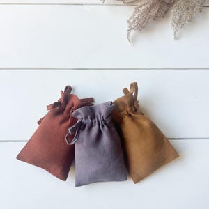 Puede incluir: Tres peque&ntilde;as bolsas con cord&oacute;n en tonos &oacute;xido, lavanda y beige est&aacute;n dispuestas sobre una superficie de madera blanca. Las bolsas est&aacute;n hechas de una tela texturizada y tienen un dise&ntilde;o simple y elegante. Hierba ornamental seca en el fondo.