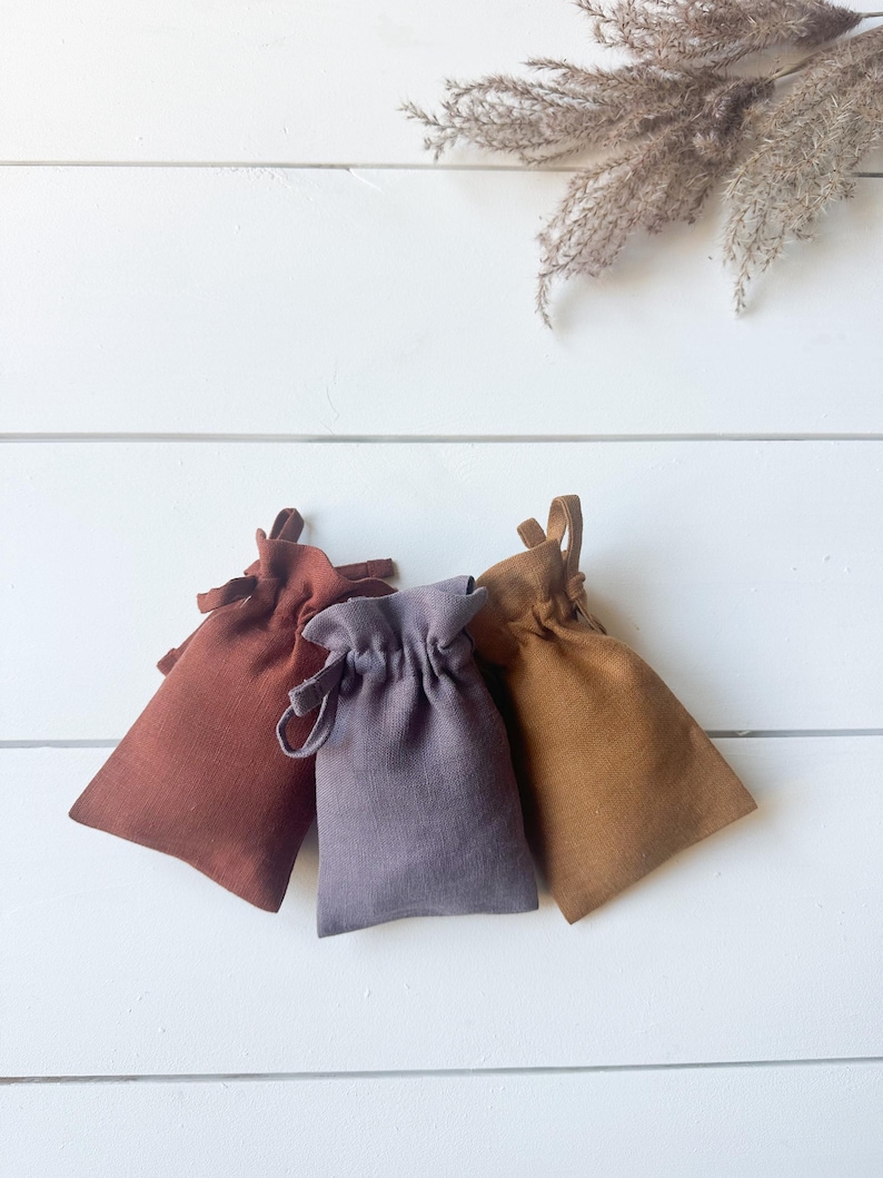 Puede incluir: Tres peque&ntilde;as bolsas con cord&oacute;n en tonos &oacute;xido, lavanda y beige est&aacute;n dispuestas sobre una superficie de madera blanca. Las bolsas est&aacute;n hechas de una tela texturizada y tienen un dise&ntilde;o simple y elegante. Hierba ornamental seca en el fondo.
