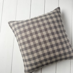 Puede incluir: Cojín cuadrado con un patrón de cuadros grises y beige. El cojín está colocado sobre una superficie de madera blanca. La tela del cojín tiene un aspecto suave y texturizado, y el patrón está distribuido uniformemente.