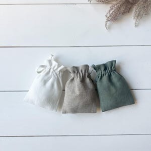 Puede incluir: Tres peque&ntilde;as bolsas con cord&oacute;n en blanco, gris y verde est&aacute;n dispuestas sobre una superficie de madera blanca. Las bolsas est&aacute;n hechas de una tela texturizada y cada una est&aacute; recogida en la parte superior con un cierre de cord&oacute;n. Hierba ornamental seca en el fondo.