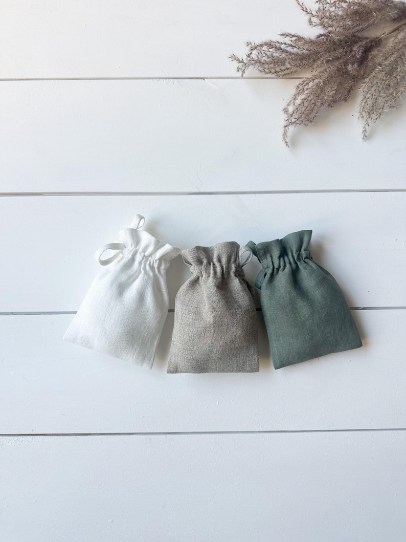 Puede incluir: Tres peque&ntilde;as bolsas con cord&oacute;n en blanco, gris y verde est&aacute;n dispuestas sobre una superficie de madera blanca. Las bolsas est&aacute;n hechas de una tela texturizada y cada una est&aacute; recogida en la parte superior con un cierre de cord&oacute;n. Hierba ornamental seca en el fondo.