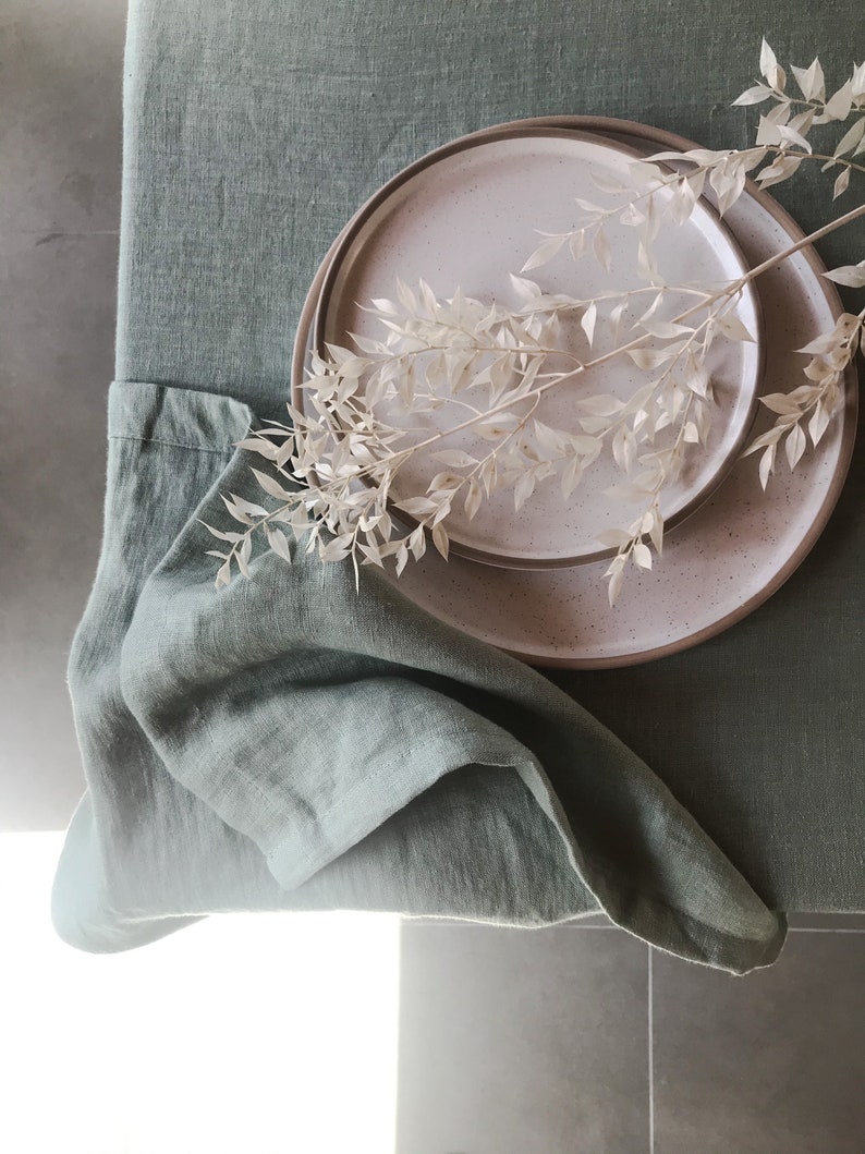 Puede incluir: Un mantel de lino verde salvia con un plato de cer&aacute;mica blanco y marr&oacute;n moteado y una rama de flores blancas secas.