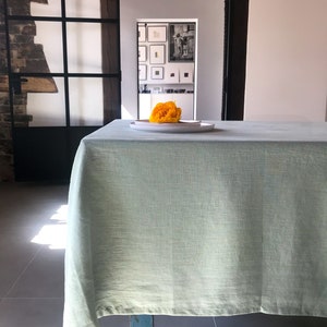 Puede incluir: Un mantel de mesa de lino verde claro drapeado sobre una mesa de madera con una sola flor amarilla en un plato blanco en el centro.
