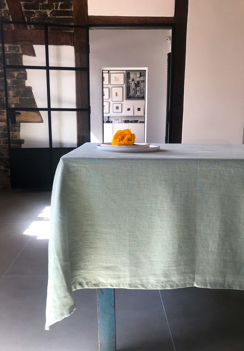 Puede incluir: Un mantel de mesa de lino verde claro drapeado sobre una mesa de madera con una sola flor amarilla en un plato blanco en el centro.