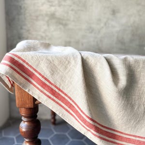 K&ouml;nnte beinhalten: Eine nat&uuml;rliche Leinen-Tischdecke mit roten Streifen, drapiert &uuml;ber einen Holztisch mit gedrehten Beinen. Die Tischdecke ist ein klassischer Landhausstil.