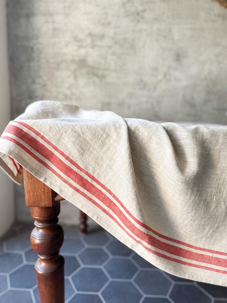 K&ouml;nnte beinhalten: Eine nat&uuml;rliche Leinen-Tischdecke mit roten Streifen, drapiert &uuml;ber einen Holztisch mit gedrehten Beinen. Die Tischdecke ist ein klassischer Landhausstil.