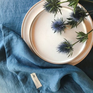 May include: A white ceramic plate with a blue linen napkin on a blue linen tablecloth. The plate is decorated with a cluster of dried thistle flowers.