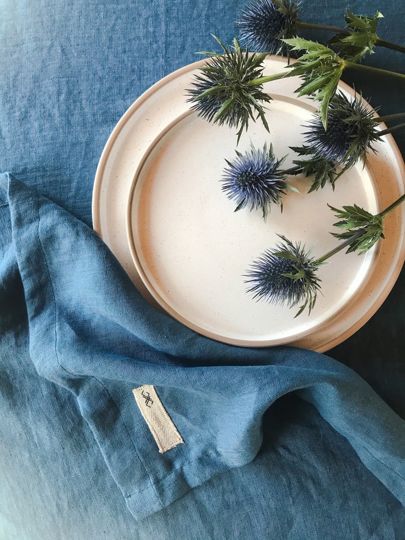 May include: A white ceramic plate with a blue linen napkin on a blue linen tablecloth. The plate is decorated with a cluster of dried thistle flowers.