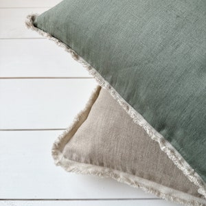 May include: Two decorative throw pillows. The top pillow is sage green, and the bottom pillow is a light beige. Both pillows have a frayed edge detail. The pillows are made of a textured fabric and rest on a white surface.