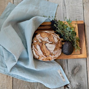 Op de afbeelding: Een lichtblauwe linnen keukendoek met een bruin label met de tekst "WENA" op een houten oppervlak met een brood, een avocado en een takje groene kruiden.