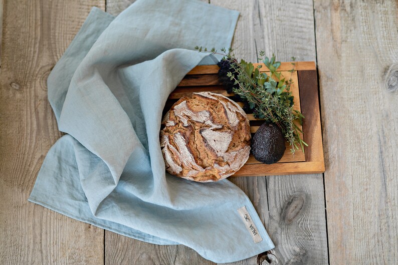 Op de afbeelding: Een lichtblauwe linnen keukendoek met een bruin label met de tekst "WENA" op een houten oppervlak met een brood, een avocado en een takje groene kruiden.