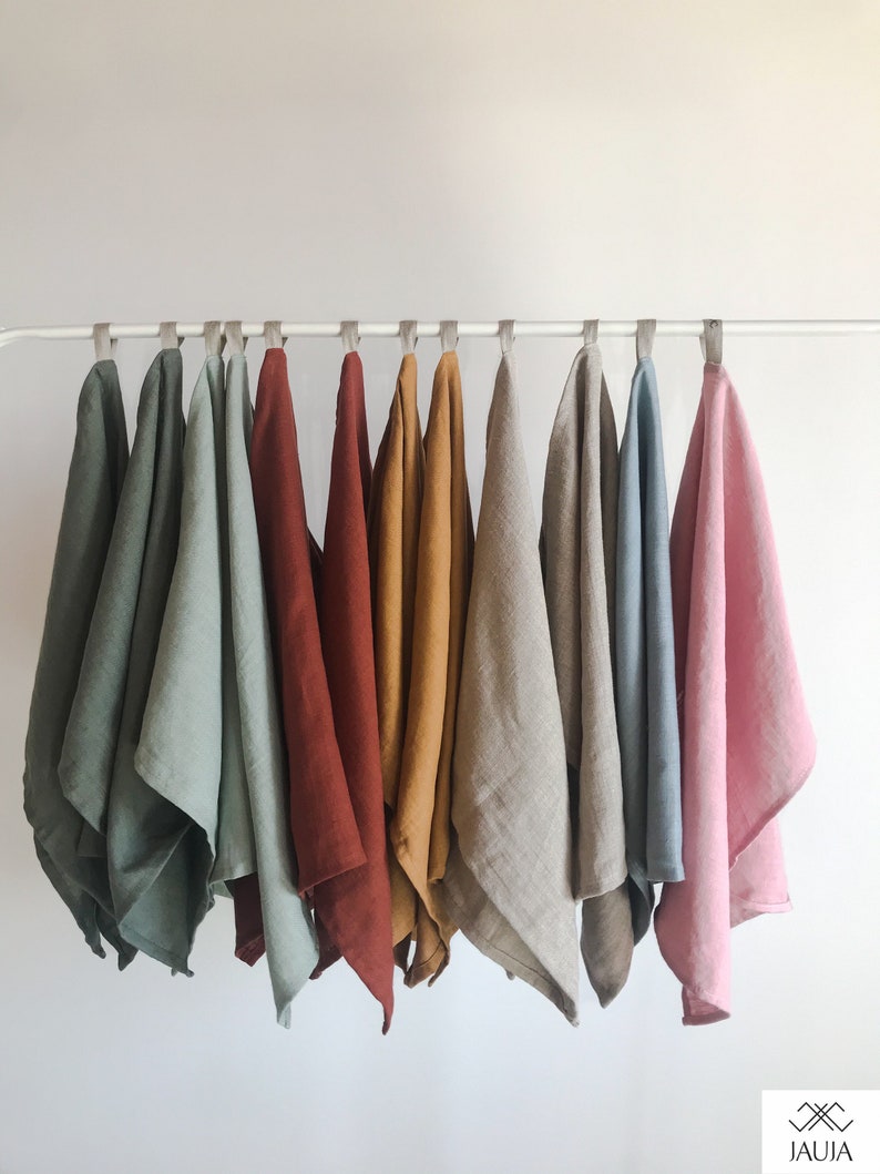 May include: A set of ten linen tea towels hanging on a white rod. The towels are in various colors, including green, red, brown, yellow, beige, blue, and pink.