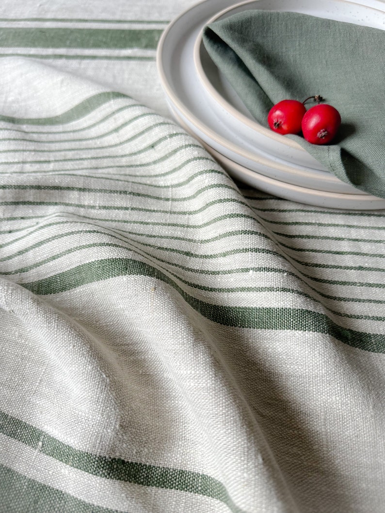 K&ouml;nnte beinhalten: Eine wei&szlig;e Tischdecke aus Leinen mit gr&uuml;nen Streifen. Die Tischdecke ist gefaltet und &uuml;ber einen Tisch gelegt. Auf einem wei&szlig;en Teller liegen zwei rote Beeren auf einer gr&uuml;nen Serviette.