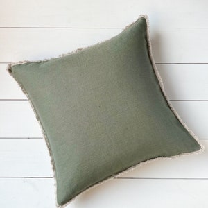 May include: Square olive green pillow with a fringed edge. The pillow has a solid color design and is placed on a white wooden surface. The pillow is a decorative home accent.