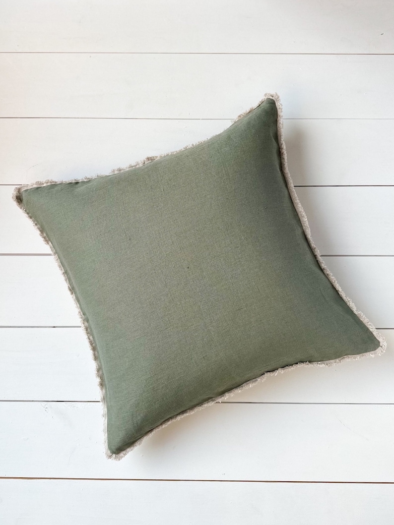 May include: Square olive green pillow with a fringed edge. The pillow has a solid color design and is placed on a white wooden surface. The pillow is a decorative home accent.