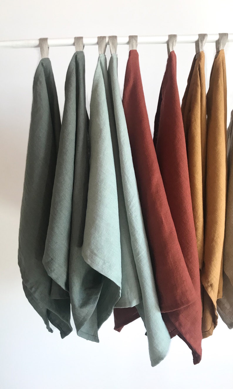 May include: Seven linen tea towels hanging on a white rod. The towels are in various shades of green, blue, red, yellow, and orange.