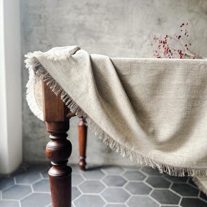 May include: A beige linen tablecloth with a fringed edge drapes over a wooden table with turned legs. The table is set on a gray hexagonal tile floor.