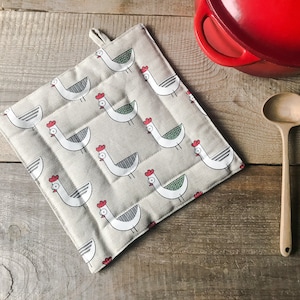Può includere: Un sottopentola in lino beige con un motivo ripetuto di galline bianche con creste rosse e accenti verdi.