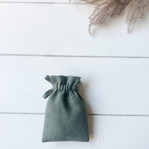 Puede incluir: Una bolsa con cord&oacute;n de lino verde salvia. La bolsa est&aacute; fruncida en la parte superior y tiene un peque&ntilde;o lazo para cerrar. El fondo es una superficie de madera blanca con hierba ornamental seca.