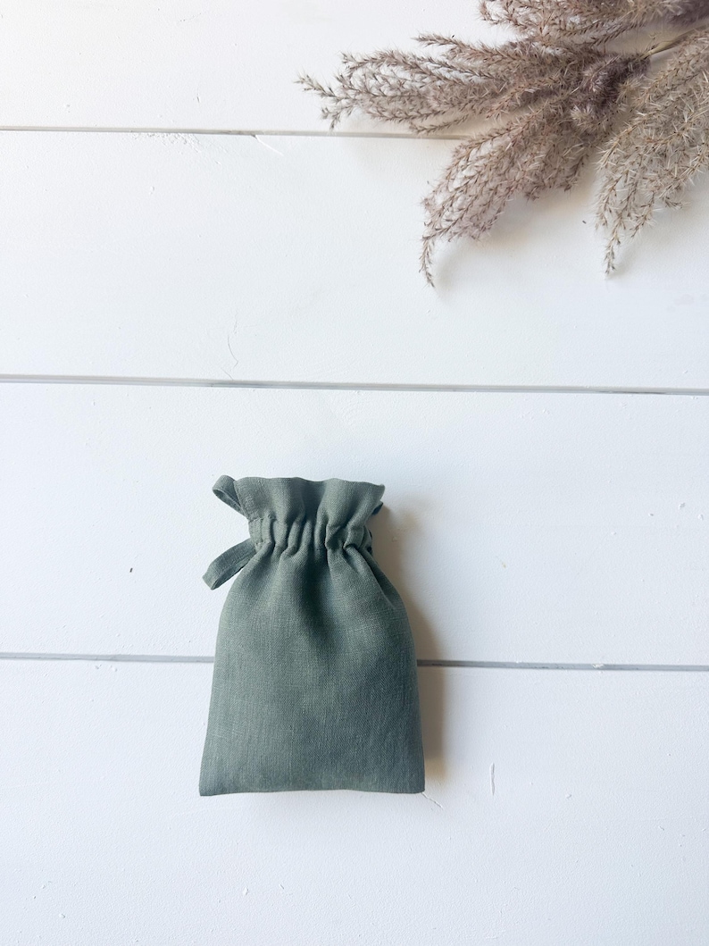 Puede incluir: Una bolsa con cord&oacute;n de lino verde salvia. La bolsa est&aacute; fruncida en la parte superior y tiene un peque&ntilde;o lazo para cerrar. El fondo es una superficie de madera blanca con hierba ornamental seca.