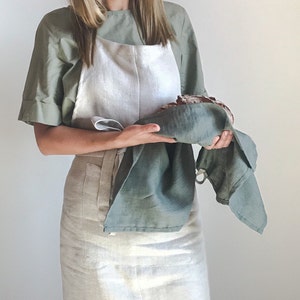 May include: A person wearing a light green shirt and a white linen apron with a green linen tea towel in their hands. The tea towel is holding a loaf of bread.
