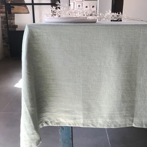 Puede incluir: Un mantel de lino verde claro drapeado sobre una mesa de madera con una sola pata visible. El mantel es liso y sin arrugas.