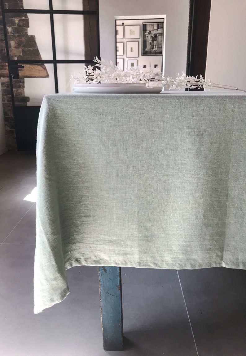Puede incluir: Un mantel de lino verde claro drapeado sobre una mesa de madera con una sola pata visible. El mantel es liso y sin arrugas.