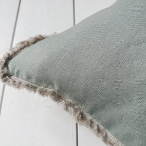 May include: Close-up of a square, sage green pillow with a textured linen appearance. The pillow has a fringed edge in a neutral, light brown color, adding a decorative element. The pillow is placed on a white wooden surface.