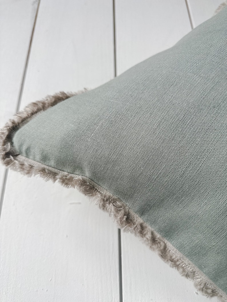 May include: Close-up of a square, sage green pillow with a textured linen appearance. The pillow has a fringed edge in a neutral, light brown color, adding a decorative element. The pillow is placed on a white wooden surface.