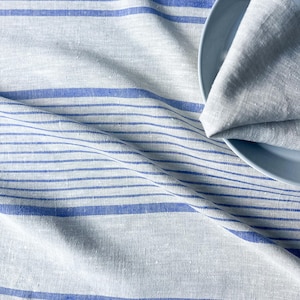 K&ouml;nnte beinhalten: Eine wei&szlig;e Tischdecke aus Leinen mit blauen Streifen. Die Tischdecke ist gefaltet und ein blauer Teller liegt darauf.