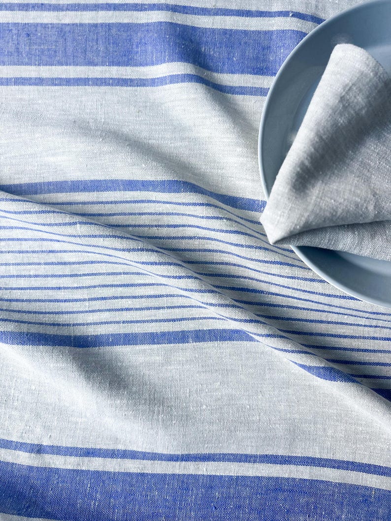 K&ouml;nnte beinhalten: Eine wei&szlig;e Tischdecke aus Leinen mit blauen Streifen. Die Tischdecke ist gefaltet und ein blauer Teller liegt darauf.