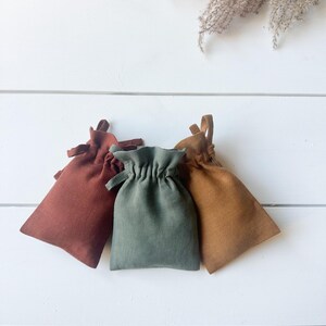 Puede incluir: Tres peque&ntilde;os bolsos con cord&oacute;n en tonos &oacute;xido, verde salvia y marr&oacute;n dorado est&aacute;n dispuestos sobre una superficie de madera blanca. Los bolsos est&aacute;n hechos de una tela texturizada y tienen un dise&ntilde;o simple y minimalista, perfecto para peque&ntilde;os regalos o almacenamiento.