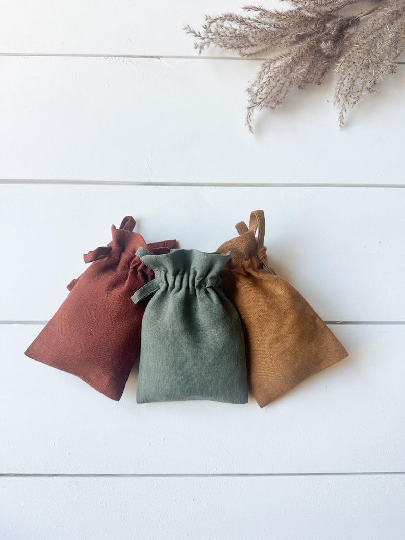 Puede incluir: Tres peque&ntilde;os bolsos con cord&oacute;n en tonos &oacute;xido, verde salvia y marr&oacute;n dorado est&aacute;n dispuestos sobre una superficie de madera blanca. Los bolsos est&aacute;n hechos de una tela texturizada y tienen un dise&ntilde;o simple y minimalista, perfecto para peque&ntilde;os regalos o almacenamiento.