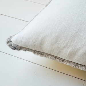May include: Close-up of a white square pillow with a textured linen fabric. The pillow has a gray fringe trim around the edges. The pillow is resting on a white wooden surface.