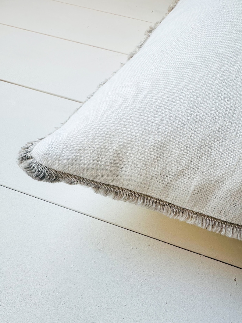 May include: Close-up of a white square pillow with a textured linen fabric. The pillow has a gray fringe trim around the edges. The pillow is resting on a white wooden surface.