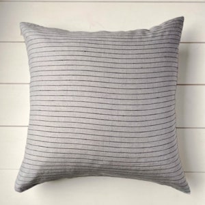 Könnte beinhalten: Ein quadratisches, graues Zierkissen mit horizontalen schwarzen Streifen. Das Kissen besteht aus einem weichen Stoff und hat ein schlichtes, modernes Design. Die neutrale Farbe würde eine Vielzahl von Einrichtungsstilen ergänzen. Ca. 40 cm x 40 cm.